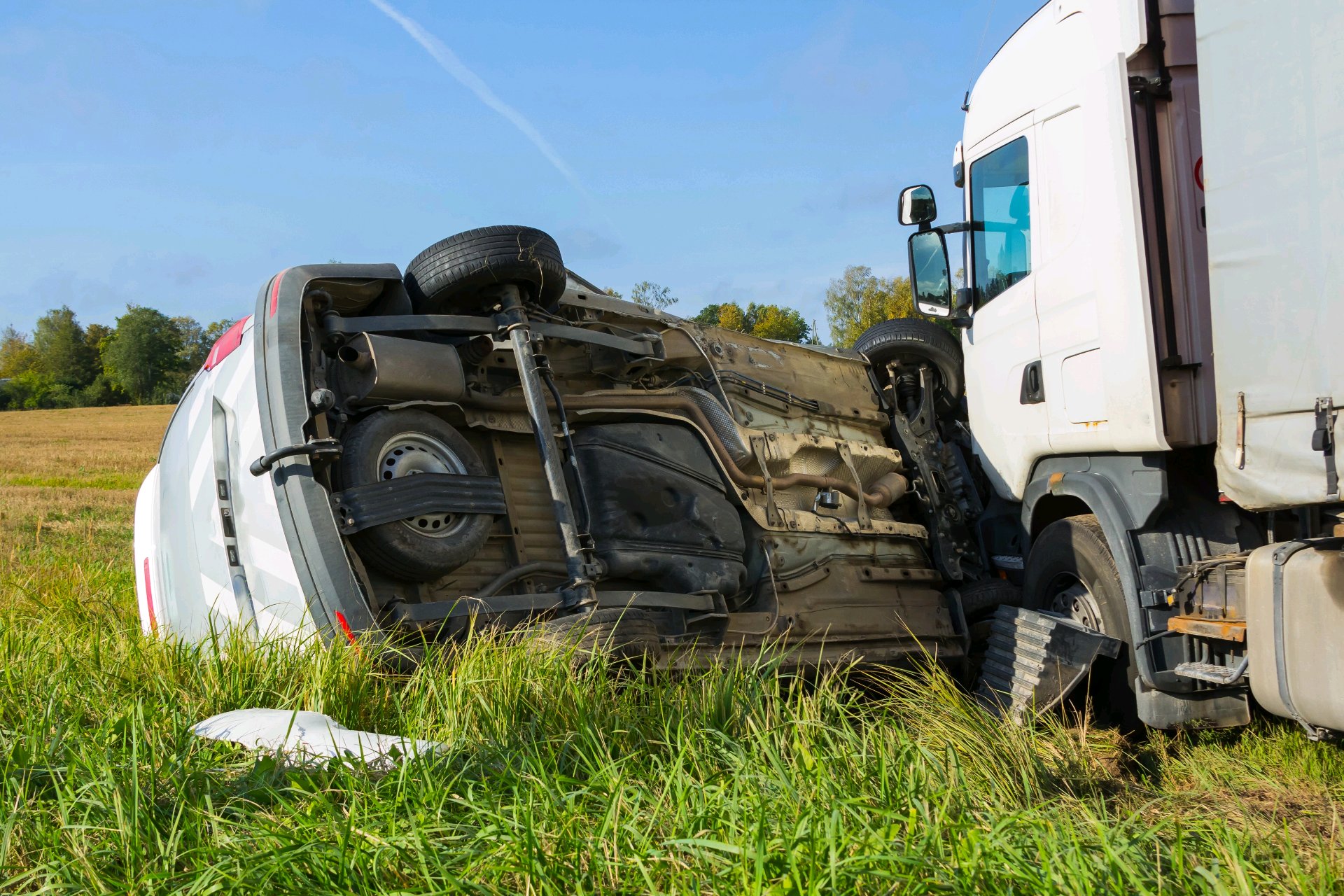 A white semi-truck rests on its side in a grassy roadside ditch beside another white truck.