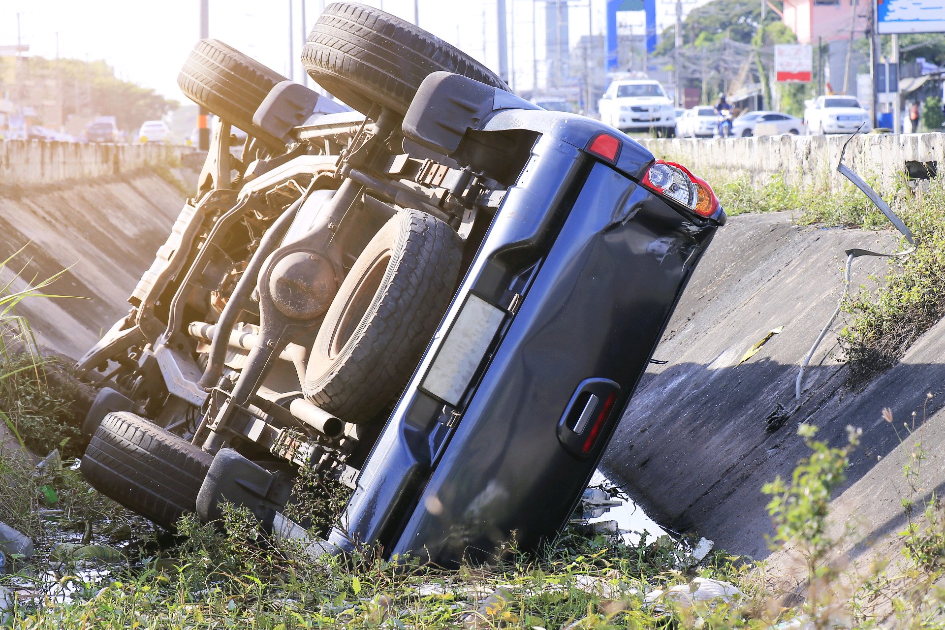 Overturned blue truck in a roadside drainage ditch beside a multilane road.
